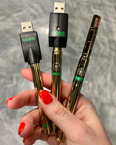 A female hand with pink nail polish holds the Ooze touchless battery, Slim Twist pen, and Slim Twist Pro in her hand above a gray floor. All three batteries are gold, and the first two have the chargers attached.