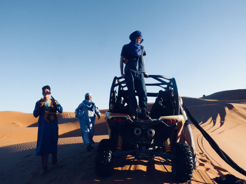 A man stands on his dune buggy in the Sahara desert. The sky is blue and the sand is orange. He is in Merzouga Morocca and by the buggy are two berber men, completely dressed in traditional outfits their suits are dyed blue with indigo
