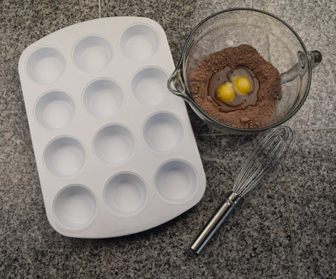 A White Ceramambake 12 cup muffin pan sits on top of a gray granite countertop, a metal whisk and a clear bowl with chocolate powder and an egg yolk in the center sit off to the side. 