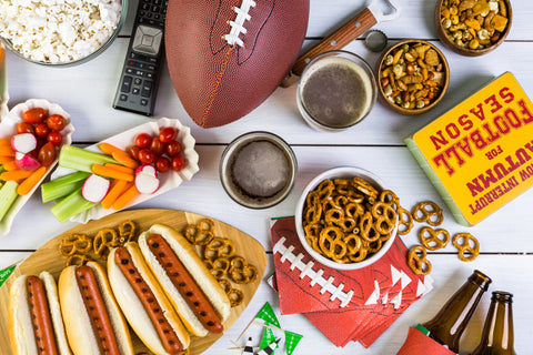 Image features a football and football themed decorations sitting on a table. The table is topped with a plate of grilled hotdogs, a plate of vegetables and drip and some amber glass bottles sit off to the side.