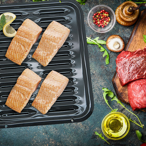 Heavy Duty Black Porcelain Range Kleen Broiler Pan features pieces of raw salmon on top of a grey background. Oil, meats and herbs are pictured on the right side of the image.