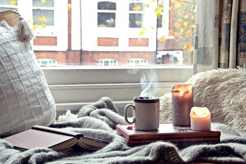 Image of steaming coffee mug with lit candles sits on wooden mat, in a cozy environment featuring blankets and pillows with a city window view as the background. An open, hardback book lays pages down nearby. 