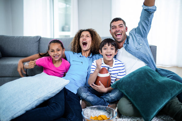 A mother, father, daughter and son sit on the couch. Arms angled up, mouths indicate that they are cheering and excited. The son is holding a football.