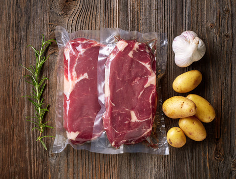 Raw steaks, in individual vacuum seal bags sit on a wooden counter top with potatoes, a garlic bulb and rosemary sprig.