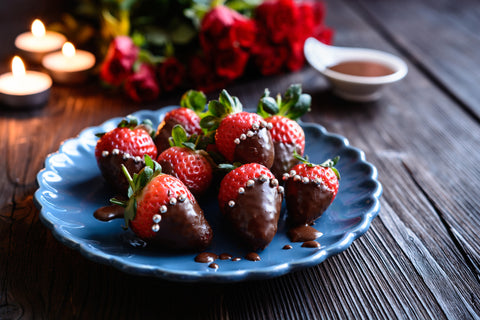Blue plate featuring fresh strawberries, half dipped in chocolate on a wooden table. Roses and votice candles appear in the background.