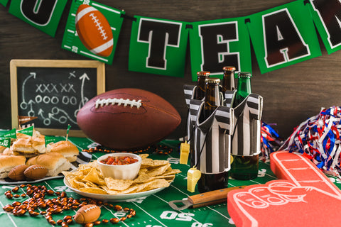 Table featuring football table cloth and various decorations, plate and bowl of chips and salsa, plate of finger sandwiches.