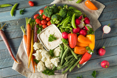 Round tray filled with raw vegetables: peppers, tomatoes, carrots, broccoli, and a container of dipping sauce on a table background.