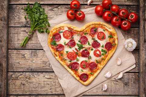 A heart shaped pizza topped with tomatoes and basil sits on pachment paper on a wooden table.