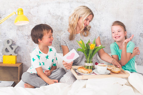 A woman with blond hair, a young boy and a young girl sit smiling on a bed with a tray of breakfast foods and flowers. 