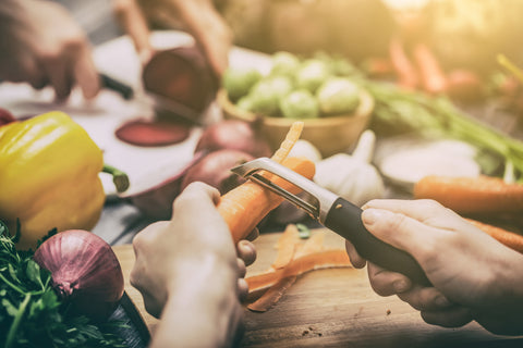 A hand holds one carror while another hand holds onto a vegetable peeler, peeling the vegetable. The background features a purple onion, yellow bell pepper, and bowl of other green vegetables.