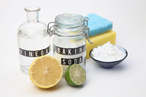 An image of clear bottles labeled Vinegar and Baking Soda with ingredients inside sit on a white background with cut lemons and limes as accents. Sponges and a bowl of baking soda sit in the background. 