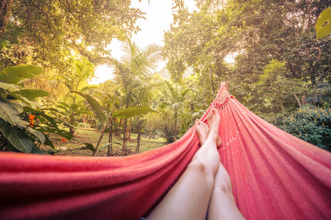 In an outdoor setting surrounded by trees, a red hammock shows the view of two human legs, with the feet crossed, relaxing.