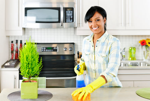 An image of a smiling woman wearing yellow cleaning gloves, holding a sponge and bottle of cleaner as she cleans a countertop. A kitchen range and sink are visible in the background.