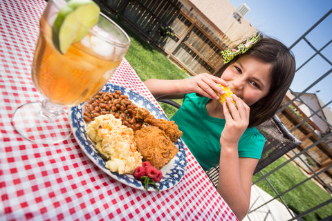 Image features girl biting into a piece of corn on the cob. The plate sitting in front of her features fried chicken, mashed potatoes and baked beans. The table is covered with a red and white checkered cloth in an outdoor setting.