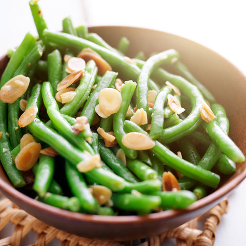 A bowl of fresh green beans topped with nuts on a table background.