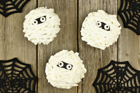 Chocolate cupcakes iced with white frosting in strips to resemble a mummy, with candy eyes in the center on a table backround.