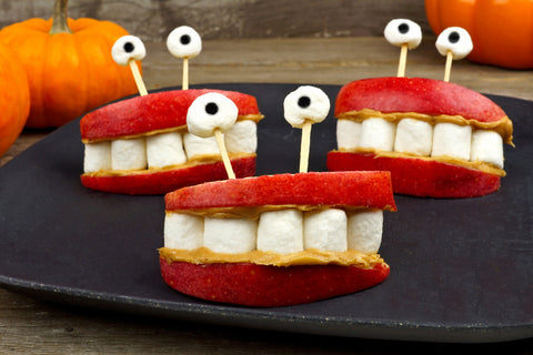 Apple mouths created using two red apple slices smeared with peanut butter, and mini marshmallows placed in the center to resemble teeth. Several sets of "mouths" sit on a dark background with toothpicks and marshmallow "eyes"