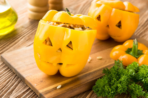 Yellow and orange bell peppers stuffed with ingreidents feature a cut out jack-o-lantern style face. Peppers sit on a cutting board in the kitchen.