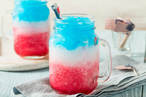 Glass mason jar mugs with handles feature a frozen drink with red, white and blue layers on a patriotic background.
