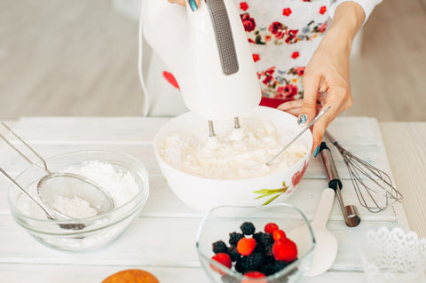A hand holds onto a hand mixer while holding a bowl of whipped cream. There is a bowl of confectioners sugar with a strainer and a bowl of assorted berries on the white countertop.