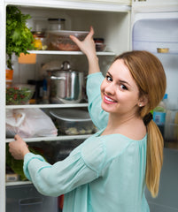 A woman reaches for a food container, smiling. The fridge is filled with food and has a white interior.