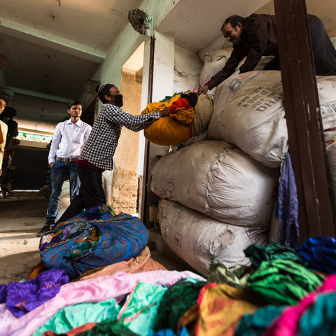 3 men are working on distributing sari fabric. One man is kneeling on a large pile of fabric bales, passing down a bale of fabric to a man standing at the bottom of the pile. Another man is standing in the doorway, waiting to be handed a bale of sari material.