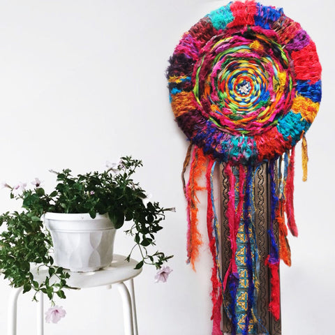 Multicolored DIY Recycled Dreamcatcher hanging on a white wall next to a white plant stand and potted house plant