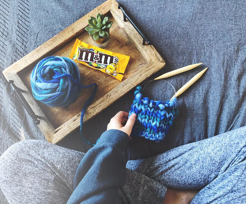 Bird's eye view of woman sitting on a bed with a wooden tray, a blue ball of yarn, circular knitting needles, and an open bag of m&ms
