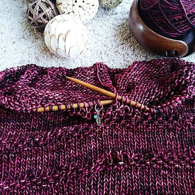 Circular knitting needles with a plum colored knitting project, yarn bowl, yarn ball, and decorative balls on a white surface