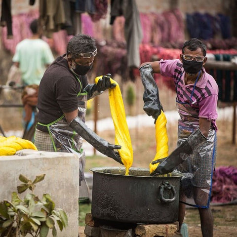 Two men standing next to a metal dye bath holding large pieces of sari silk ribbon yarn in yellow, and wearing aprons and masks