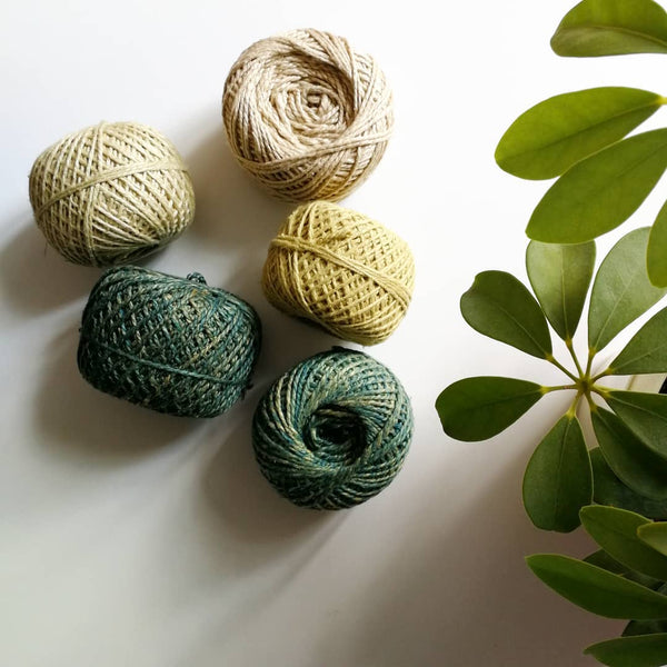 Multiple skeins of herbally dyed yarn in different ombre shades of green. To the right of the image is a leafy house plant.