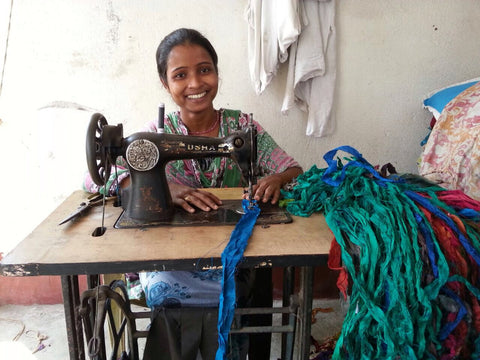 An Indian artisan seated at a sewing machine, working on a pile of torn sari fabric and smiling at the camera