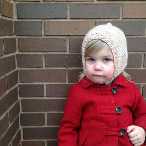 Little girl wearing a cream Merino Wool Baby Hood and a red coat and standing in front of a brick wal