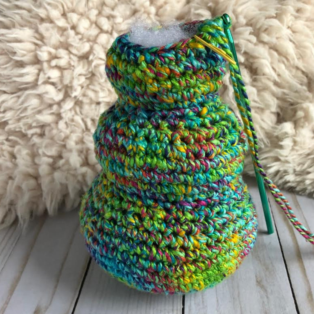 Partially crocheted blue and green Christmas Tree with a green crochet hook sitting on a wooden surface next to a white fur blanket