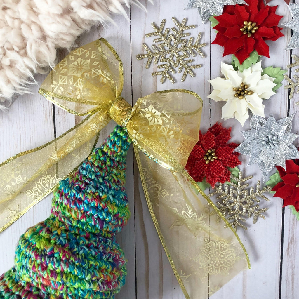 Blue and green crochet Christmas Tree with a gold bow on top sitting on a wooden surface next to faux poinsettia flowers, snowflakes, and a fur blanket