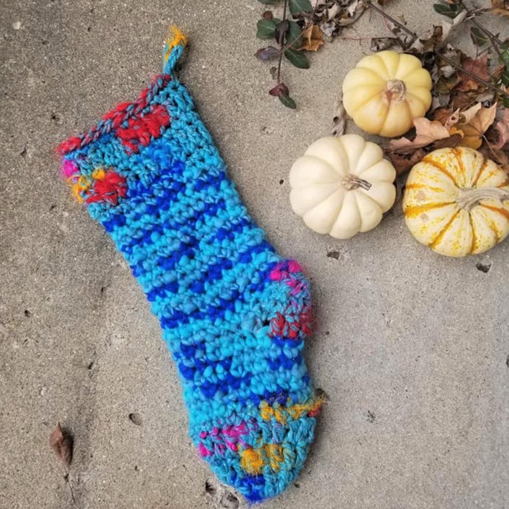 Blue Crochet Christmas Stocking sitting on concrete surface next to three pumpkins and fall leaves