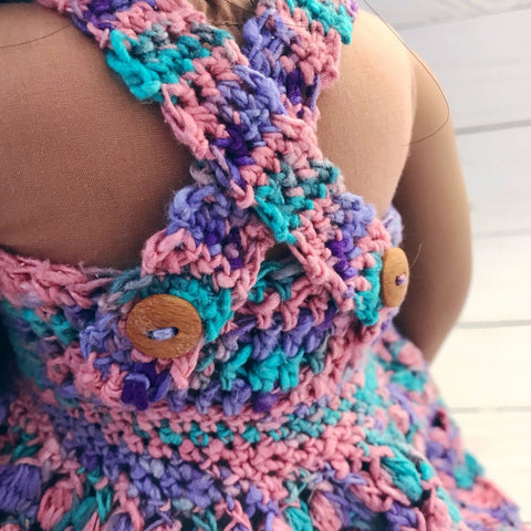 Backview of an 18" doll wearing a pink, purple, and blue crochet sundress standing in front of a white background