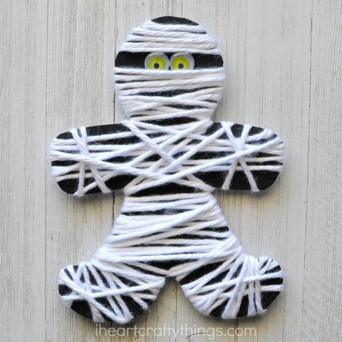Paper and yarn mummy with googly eyes sitting on a wooden surface