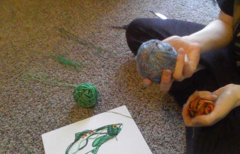 A person is sitting on the carpet. In front of them is a canvas board with a salmon drawn on it. They are holding a ball of yarn, displaying the yarn that they will glue onto the canvas board.