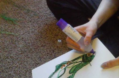 While sitting on the ground, the person is now using a glue-pen to glue the yarn onto the canvas board. The glue is placed in specific areas to create the pattern needed for the yarn painting.