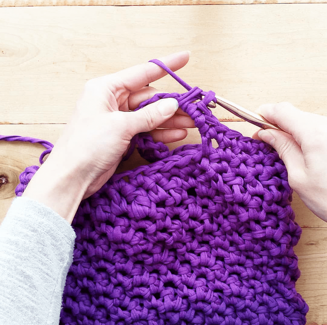 Woman's hands holding a crochet hook and purple crochet project over a wooden surface