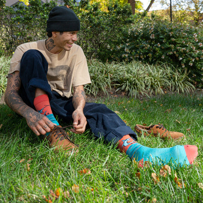 Man seated in grass taking off dress shoes, revealing the Tailwalker swordfish socks in teal blue
