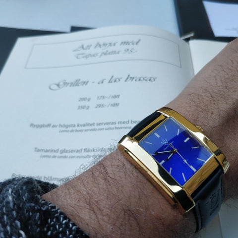 Fyrkantig herrklocka i polerat guld med blå klassisk urtavla - fyrkantig klocka herr i närbild över en restaurangmeny