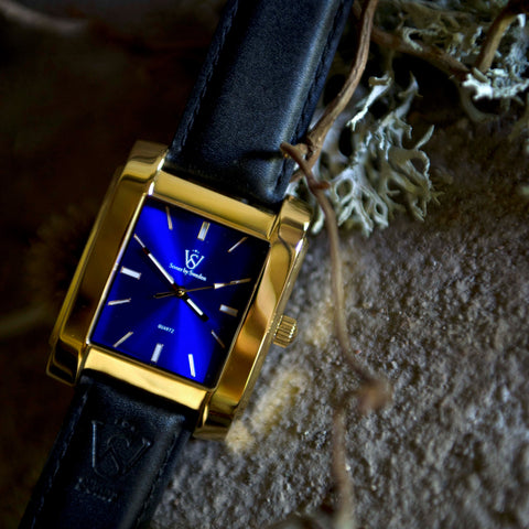 Herrlocka i polerat guld med blå klassisk urtavla - Fyrkantig klocka i närbild liggandes på en sten