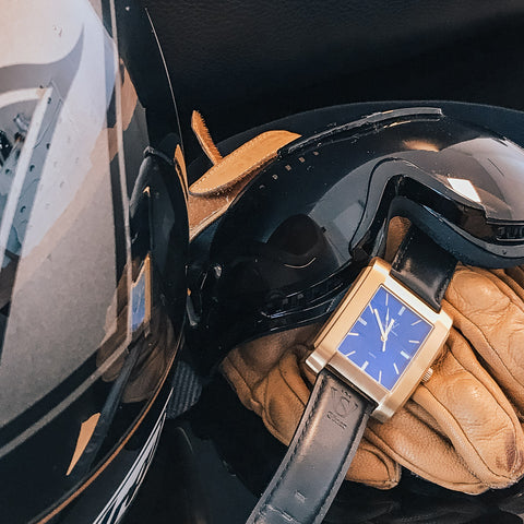 Fyrkantig herrklocka i borstat guld med blå klassisk urtavla - klocka ligger vid motorcykelhjälm, handskar och solglasögon
