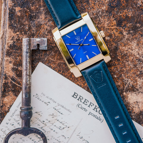 Herrklocka i polerat guld med blå klassisk urtavla - närbild på fyrkantig klocka som ligger bredvid ett gammalt vykort och en gammal nyckel