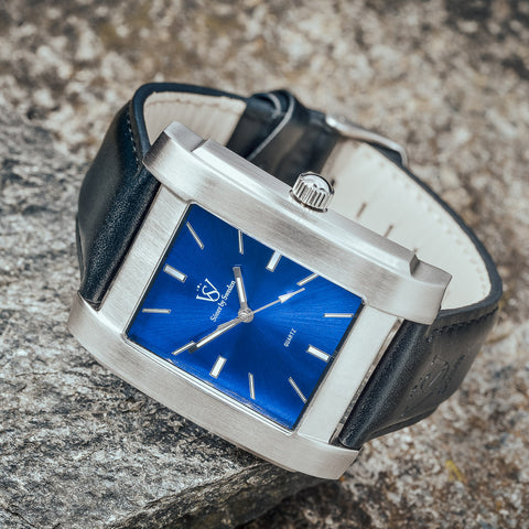 Fyrkantig herrklocka i borstat stål med blå klassisk urtavla - närbild på fyrkantig klocka som ligger på en grå sten