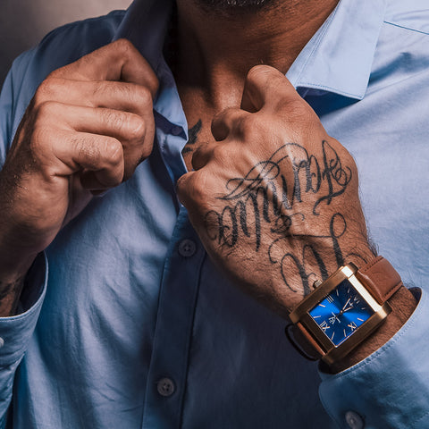 Fyrkantig herrklocka i borstat guld med blå romersk urtavla - fyrkantig klocka på tatuerad hand sträcker skjortkrage