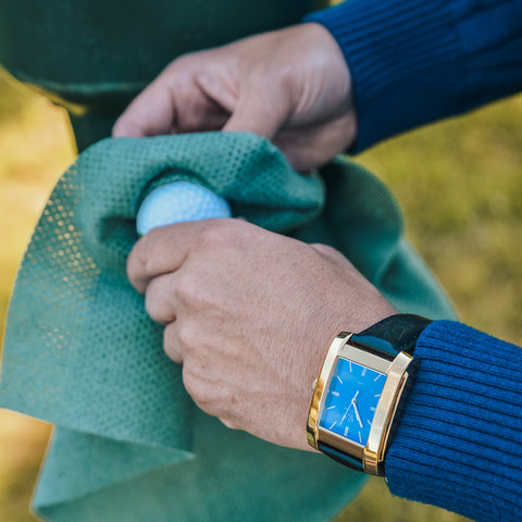Fyrkantig herrklocka i polerat guld med blå klassisk urtavla - närbild på fyrkantig klocka medans man tvättar golfboll