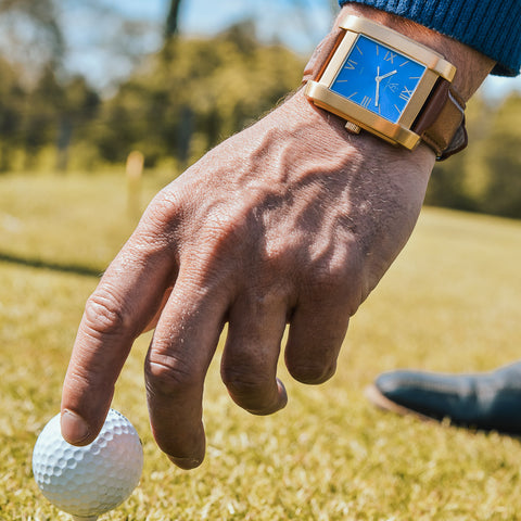 Herrklocka i borstat guld med blå romersk urtavla - närbild på fyrkantig klocka medans man peggar upp golfboll